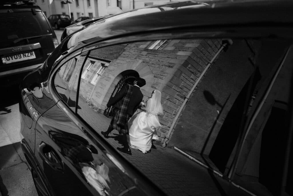 A bride and groom's reflection in a car window as they walk through St Andrews