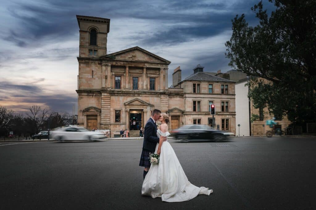 Bride and groom kiss in the middle of the street as blurred cars rush past them in Glasgow's city centre