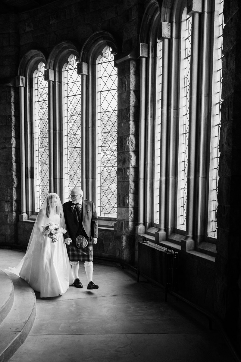 Father of the bride walks with his daughter by tall stained-glass windows, in black and white