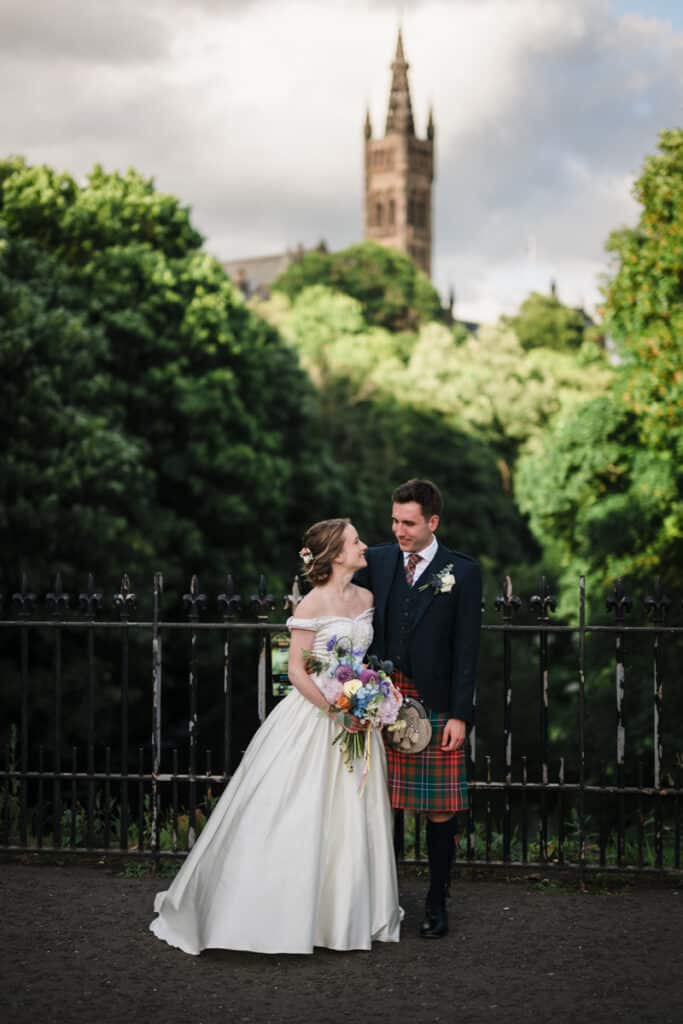 Bride and groom stand next to each other in Kelvingrove with the University of Glasgow in the background