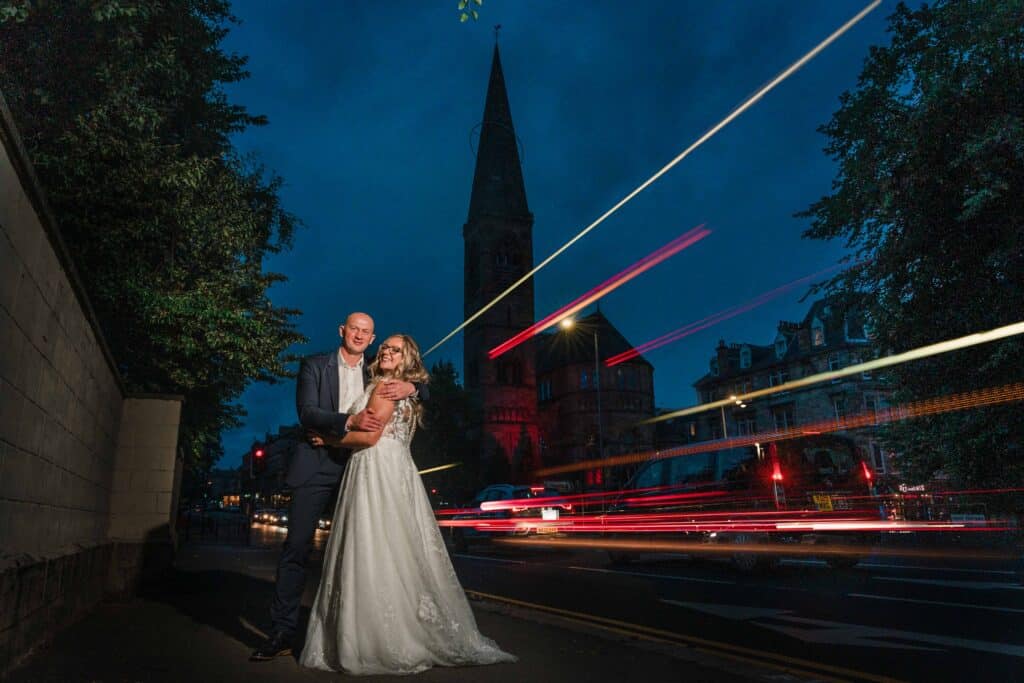 Night outside Oran Mor in Glasgow, a bride and groom stand whilst light trails from passing traffic surround them
