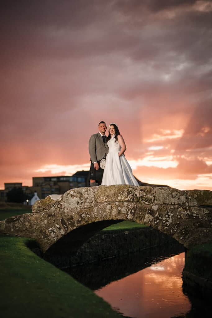 As the sun sets, a bride and groom stand on the Swilcan Bridge with the Old Course Hotel behind them