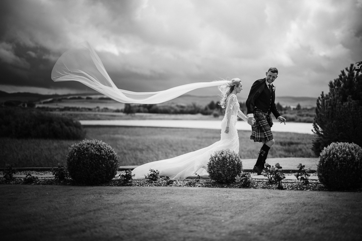 Black and White image of a bride and groom walking, with her veil flying in the wind, at Lochside House Hotel in Ayrshire