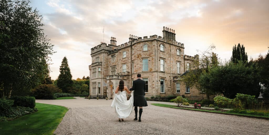 A bride and groom walk towards Crossbasket Castle during their wedding