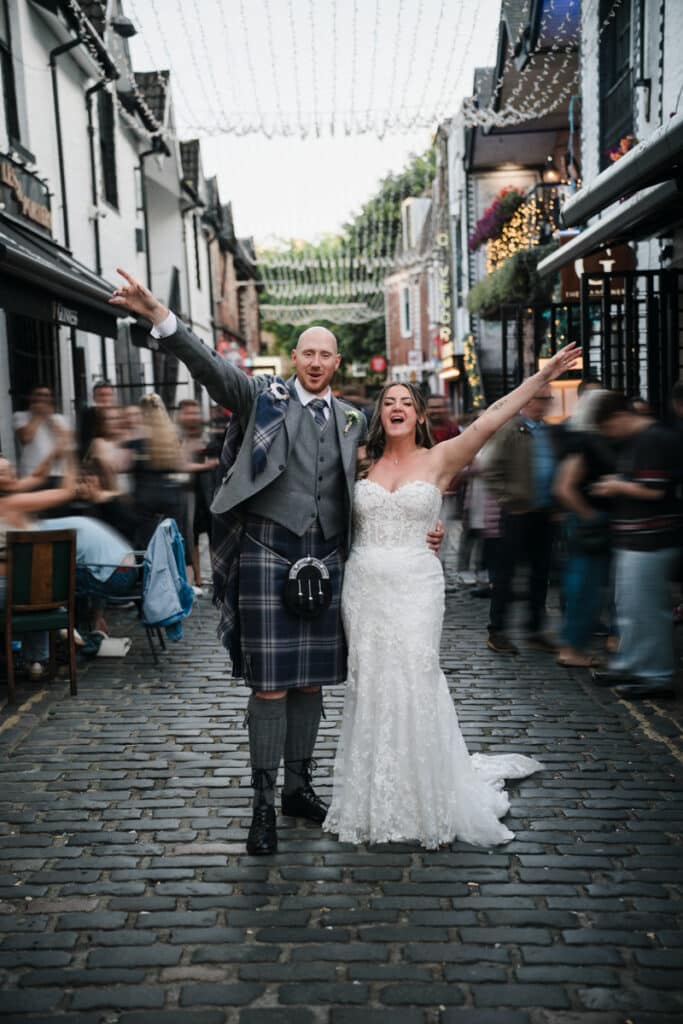 Bride and Groom stand celebrating in Ashton Lane whilst people rush past them